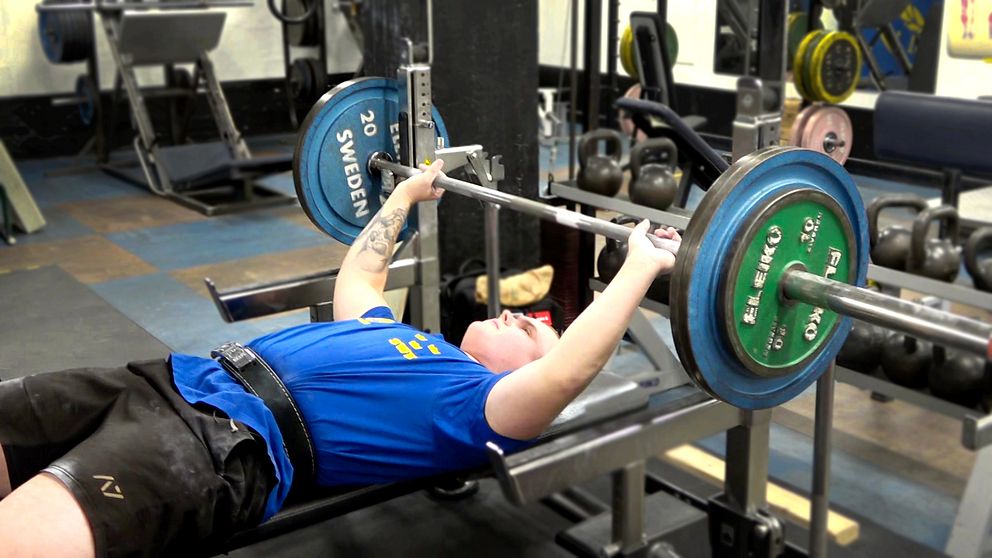 Simon Blom utför bänkpress i ett gym i Södertälje, liggande på en bänkbänk med en skivstång över bröstet. han bär en blå t-shirt med gul text och har ett bälte runt midjan för stöd. Vikterna på skivstången är markerade med färgade plattor. I bakgrunden syns andra träningsutrustningar och vikter.