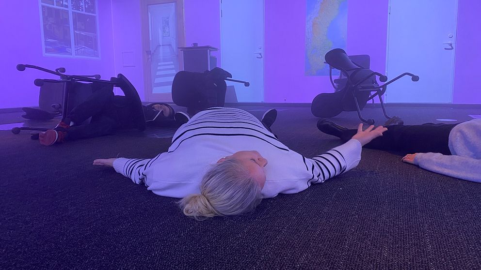 Kvinna liggande på golvet i ett kontorsmiljö med utspridda stolar och lila belysning, vilket kan indikera en simulering av en nödsituation eller träning i evakuering.