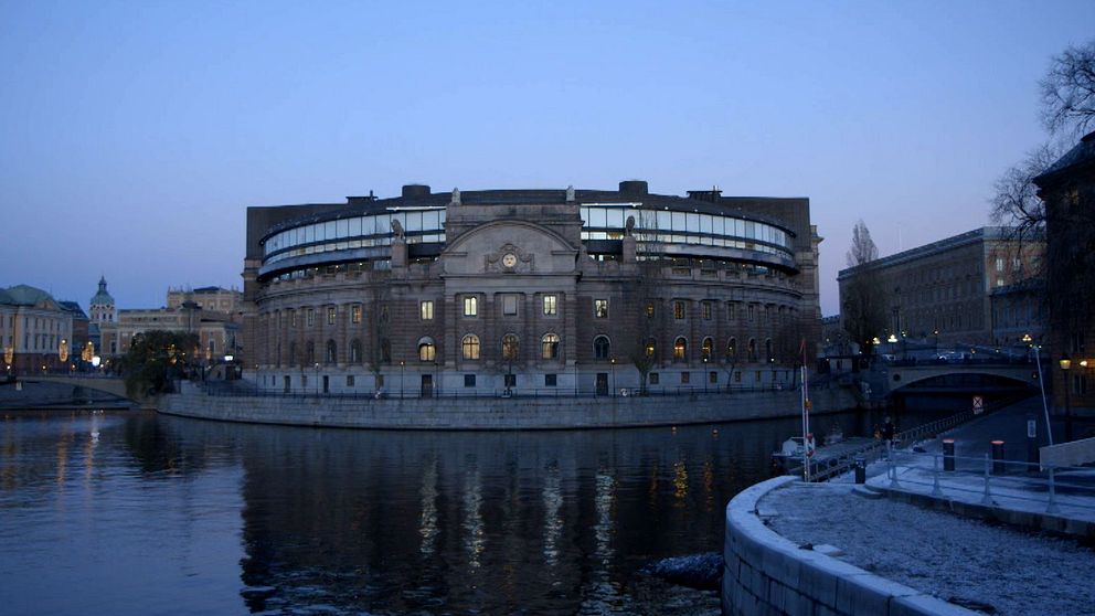 Svenkska riksdagshuset vid skymning, speglat i vattnet. Omgivningen visar en lugn stadsbild med belysning från byggnaderna i bakgrunden och en klar himmel.