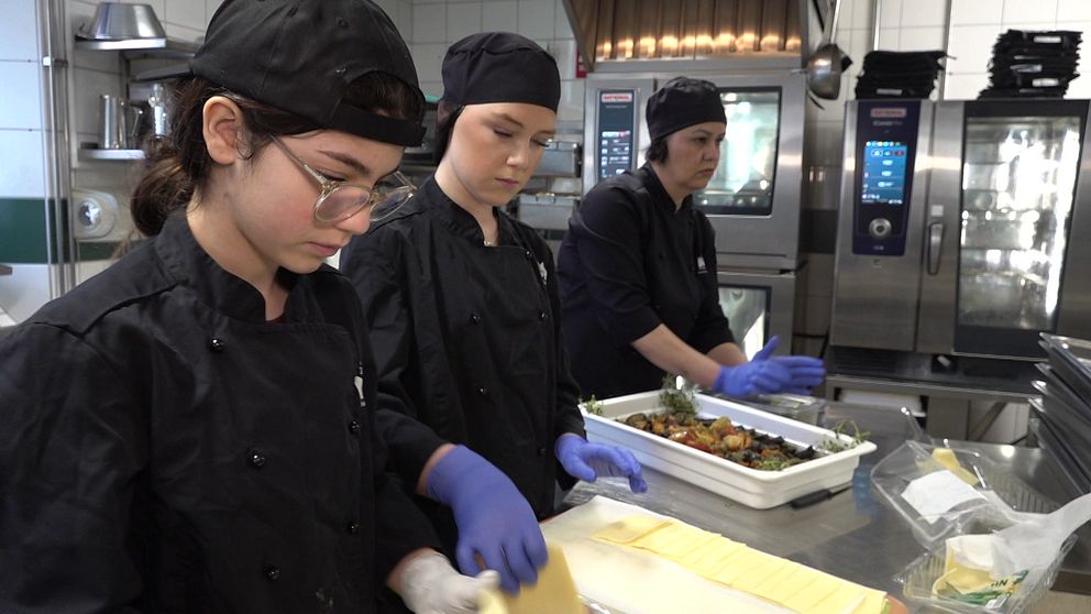Tre personer i köksuniform förbereder mat i ett skolkök.