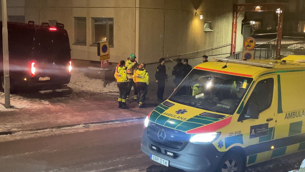 Räddningstjänst och ambulanspersonal arbetar i ett vinterlandskap vid en byggnad. En ambulans står parkerad på gatan, medan flera personer i skyddskläder diskuterar vid en svart minibus. Det är mörkt, och snö ligger på marken.
