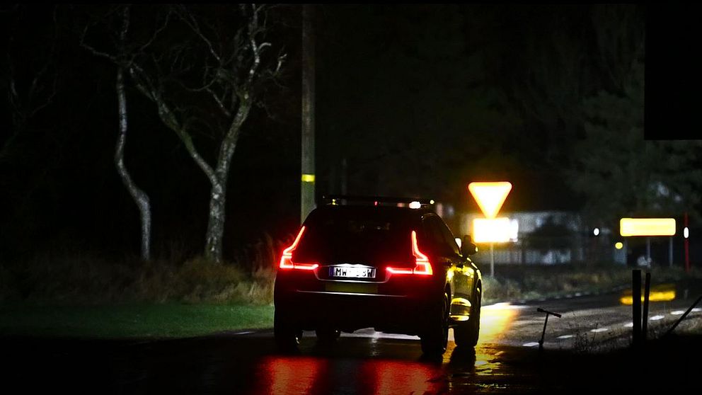 Polisbil med ljusen på kör bort från kameran på en mörk väg, med träd och vägskyltar synliga i bakgrunden. Belysningen från bilens bakljus och vägskyltarna skapar kontrast mot den nattliga miljön.