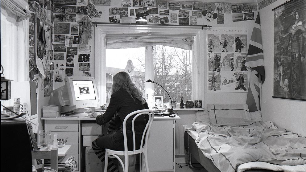 En svartvit bild där en ung kvinna sitter vid ett skrivbord. Väggarna är täckta av foton, affischer och flaggor.