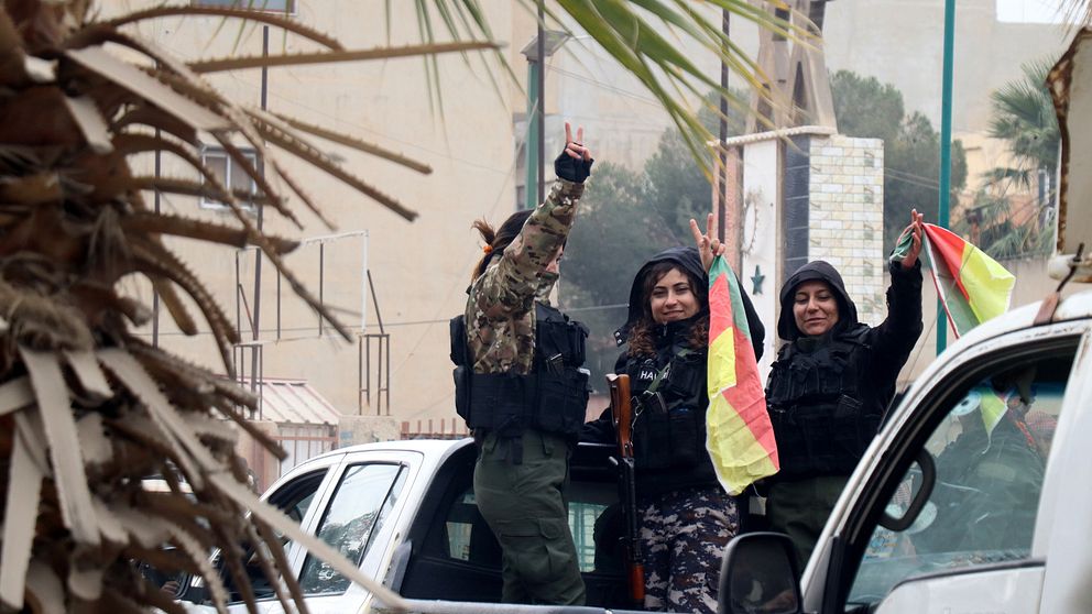 Tre kvinnliga SDF soldater med flaggor står på dlaket på en vit pickupp. De firar efter att kurdiskledda SDF intagit staden al- Hasakah i norra Syrien.