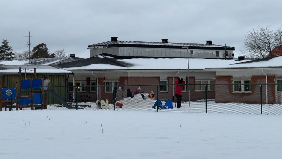 Barn leker utomhus i snön vid en förskola. I förgrunden syns ett lekställe med klätterställning och en snöhög där barn bygger. Byggnaden i bakgrunden är lågt belägen med takfönster och mörka fönster. Grå himmel och snö täcker marken.