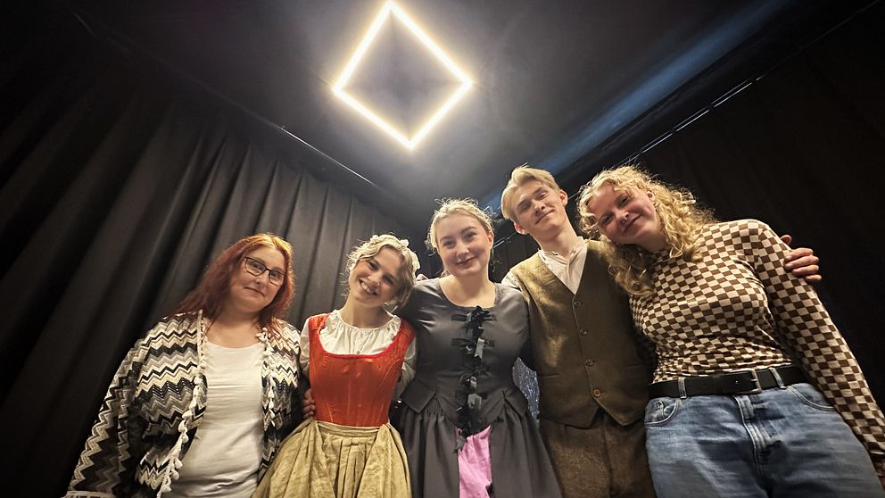 Fem personer i kostym under en ljuskub på scenen, glatt poserande inför en teateruppsättning.