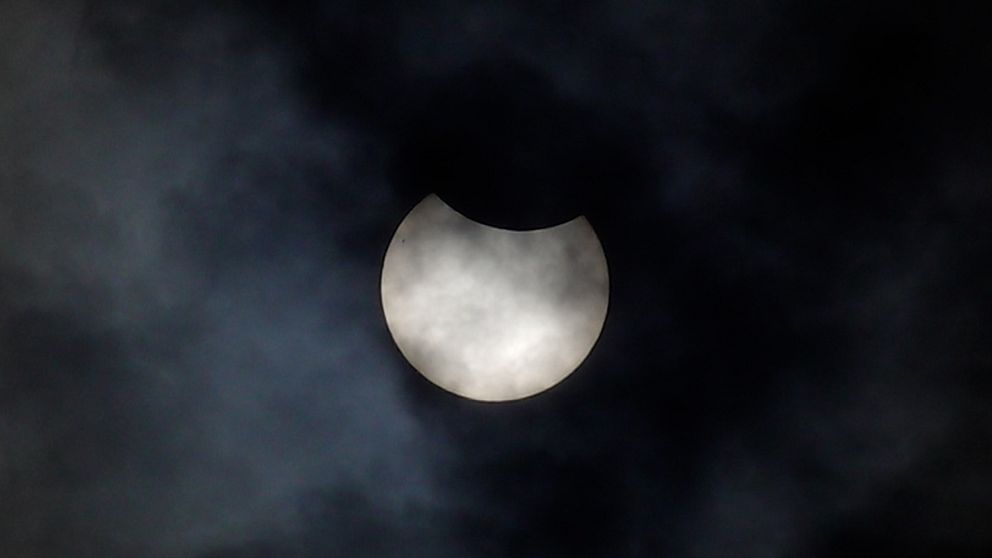 Solförmörkelse delvis dold bakom moln på mörk himmel.