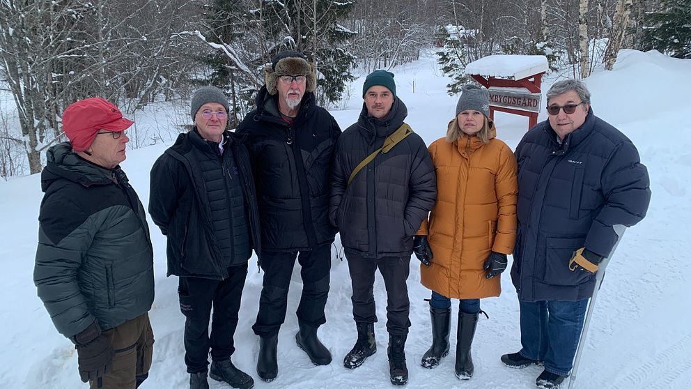 Grupp av personer i vinterkläder står på en snötäckt väg med träd i bakgrunden. En person bär en orange jacka och de andra har på sig mörka ytterkläder. I bakgrunden syns en skylt som kan indikera en väg eller plats.