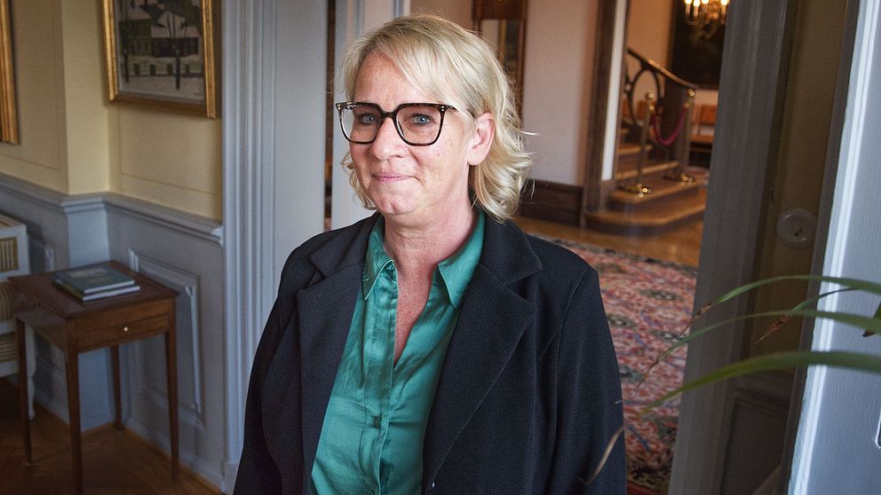 Person med ljusblondt hår i formellt rum, väggkonst och möbler synliga.