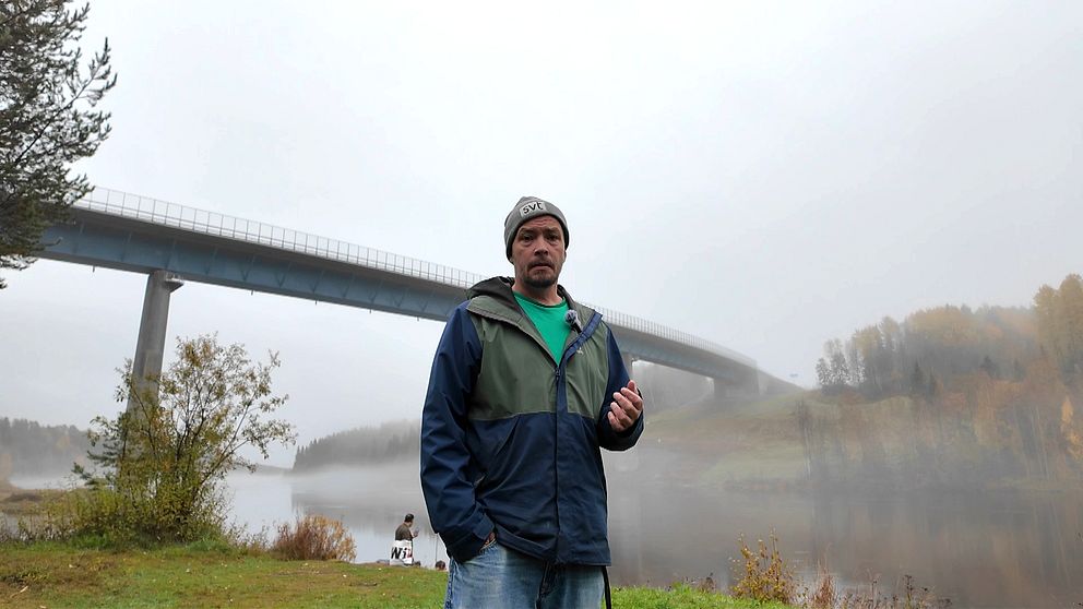 En man står vid vattnet i en dimmig miljö, med en bro som sträcker sig över en lugn älv i bakgrunden. Området omges av träd med höstliga färger, och en annan person kan ses fiska längre bort vid vattenkanten.
