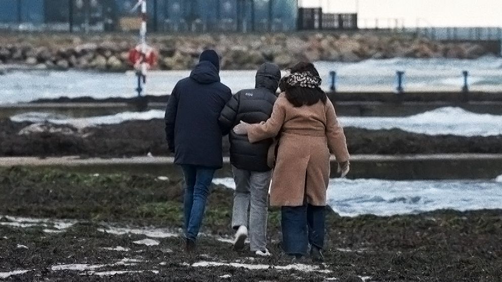Tre personer promenerar längs en kustlinje vid havet, insvepta i vinterkläder. I bakgrunden syns en byggnad nära vattnet och vågor slår mot stranden.