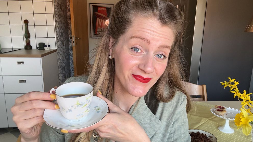 Kvinna i 1940-talsklänning och rött läppstift håller en kopp kaffe i ett kök med blommor och bakverk på bordet.