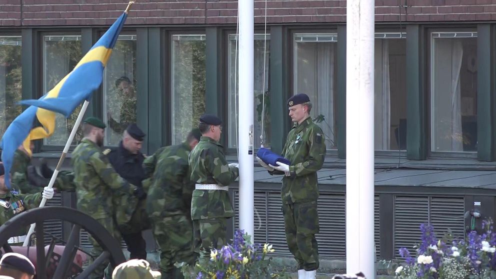 Två militärer hissar Nato-flagga, bakom dem kommer fem personer bärande på man i militärkläder.