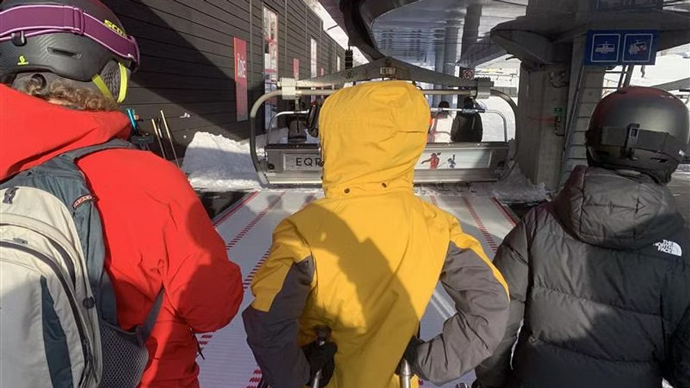 Tre personer i skidutrustning väntar vid en skidlift på en skidort. De bär hjälmar och ryggsäckar medan de står på en övergångsplatta framför liften, som är redo att ta dem uppför berget.