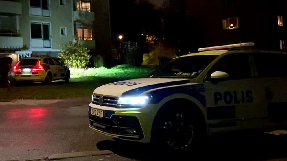 En polisbil står invid en gata och på andra sidan gatan står en annan polisbil invid ett hyreshus.