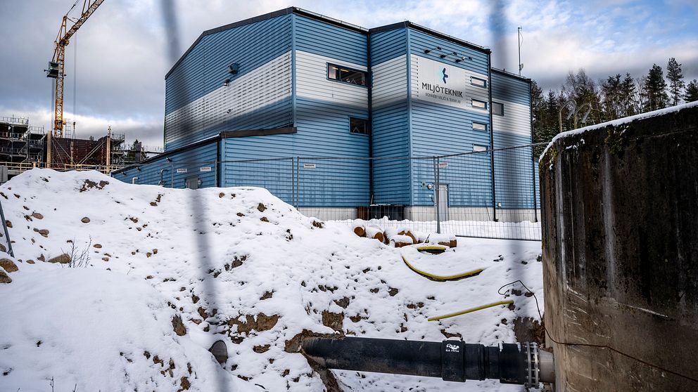Snötäckt byggarbetsplats med ett blått och silverfärgat industribyggnad, märkt med ”PilotTremin”, synlig i bakgrunden. En stor kran reser sig till vänster och en svart stor rörledning sträcker sig över snön, omgiven av byggstängsel.