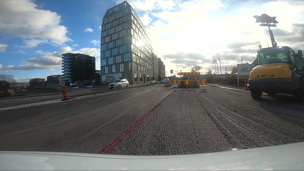 En vägscen som visar pågående vägbygge. I förgrunden syns avstängningskoner och byggutrustning, inklusive en grön maskin. Bakgrunden har en modern byggnad och några mindre byggnader. Himlen är delvis molnig med solstrålar som syns.