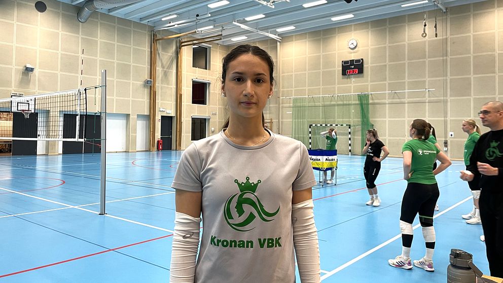 En ung kvinnlig volleybollspelare från Kronan VBK står i borstahusens idrottshall. Tränare och andra volleybollspelare syns i bakgrunden.