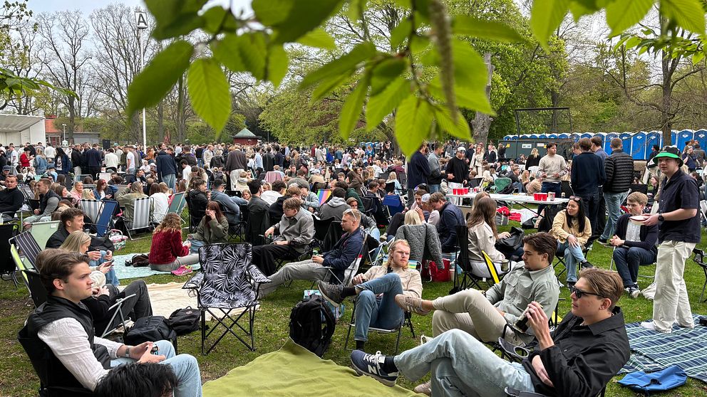 Många människor sitter på filtar och stolar i park under utomhusevenemang.