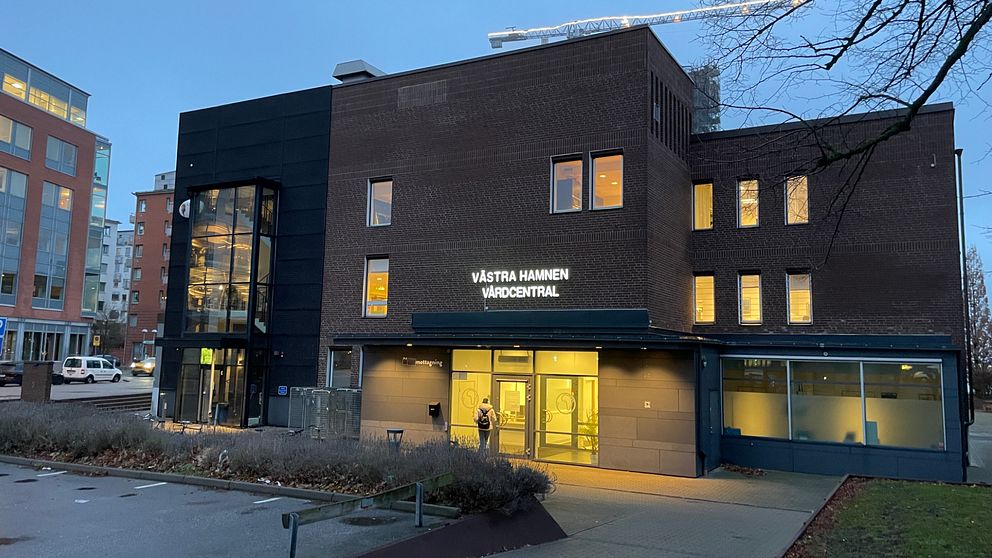 Byggnaden avbildar Västra Hamnen Vårdcentral, som har en modern design med stora fönster som lyser upp fasaden i skymningen. Entrén är välkomnande med en glasdörr och breda steg som leder in. Området är omgivet av grönområden och närliggande byggnader.