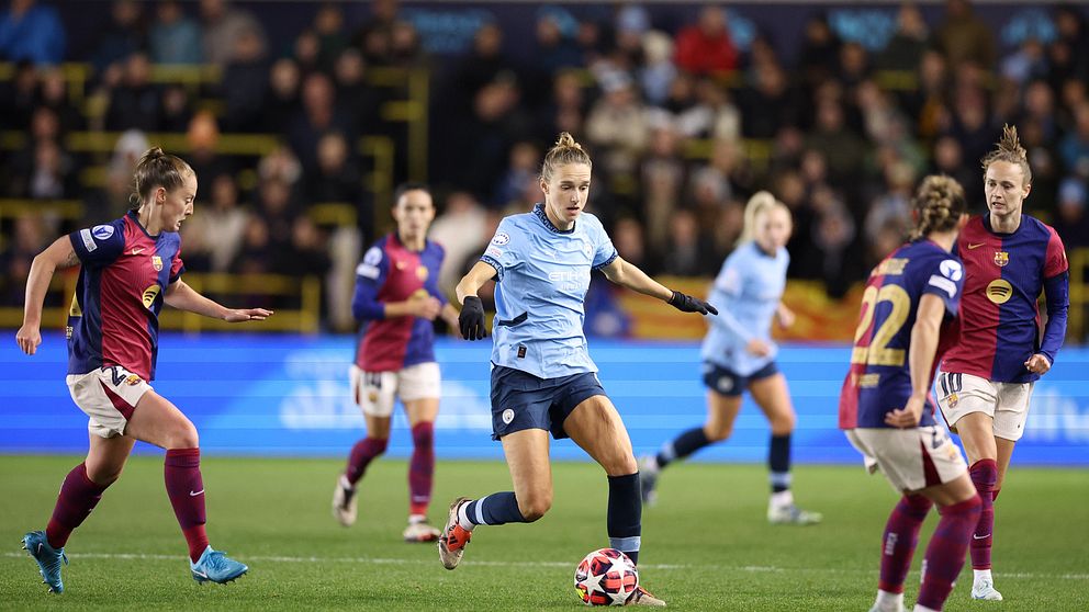 En fotbollsspelare i en ljusblå tröja dribblar bollen på planen, omgiven av motståndare i mörkröda tröjor. Bakgrunden visar en åskådarmassa samt en stor LED-skärm. Matchen verkar vara en del av en internationell tävling.