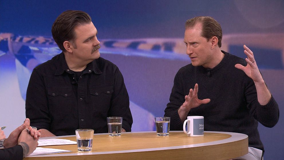 Två personer sitter vid ett runt bord i en TV-studio under ett samtal. Den ena personen gestikulerar med handen, medan den andra lyssnar uppmärksamt. På bordet finns flera glas med vatten och en mugg. En abstrakt, blå bakgrund syns bakom dem som skapar en professionell atmosfär.
