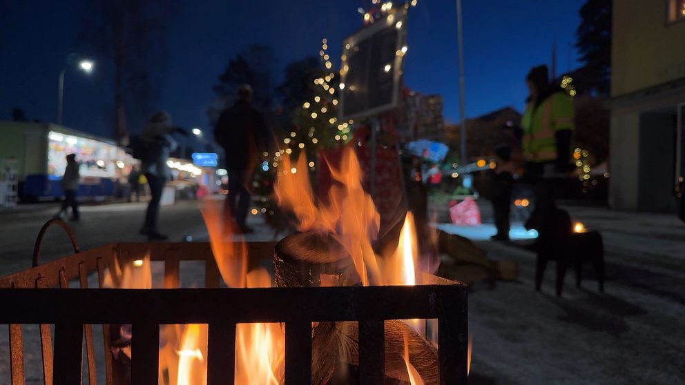 Eld i en metallbrasa i förgrunden, med en festlig gata i Storfors i bakgrunden som är belyst av dekorationer och juliga ljus. Människor rör sig runt i en vinterlig miljö under kvällens blå skymning.