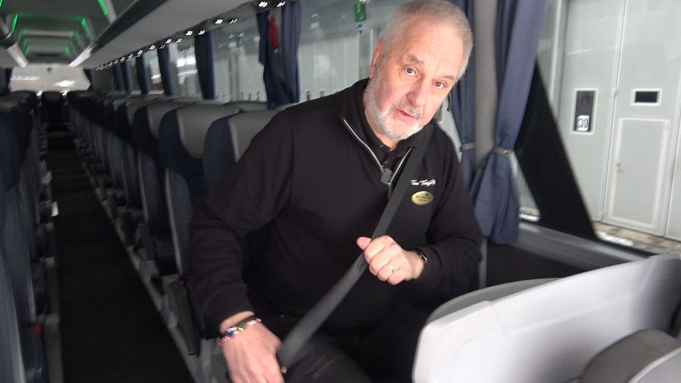 Busschauffören Stefan Wahlén sitter i en buss och fäster säkerhetsbältet, omgiven av tomma säten. Bussens interiör är modern med grå och blå klädslar och belysning i taket.