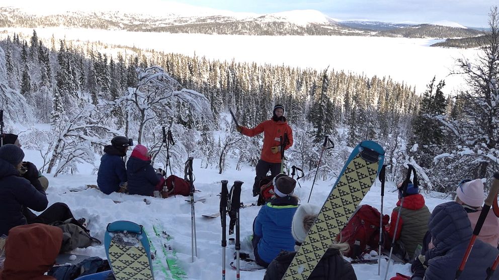 En grupp skidåkare sitter i snön och lyssnar på en guide som pratar framför dem, med en snöklädd skog och berg i bakgrunden. Vintrig landskapssceneri med skidutrustning synlig omkring dem.