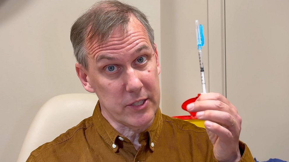 Vetenskapsreporter Johan Bergendorff håller en spruta med blått lock i en klinisk miljö.