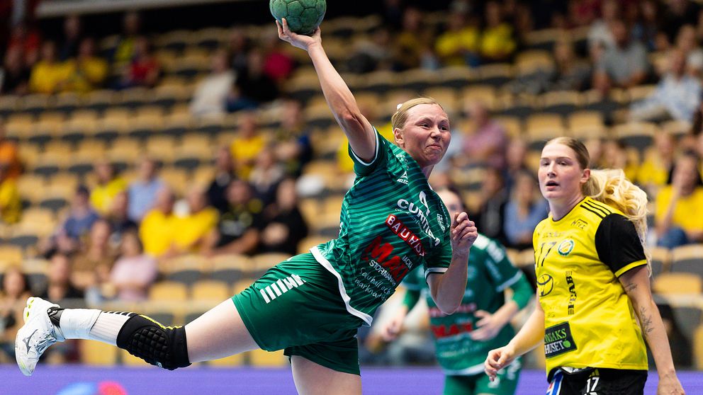 En handbollsspelare i grön tröja och svart shorts kastar bollen mot mål under en match, medan en motståndare i gul tröja observerar från sidan. Publiken i bakgrunden är delvis synlig.
