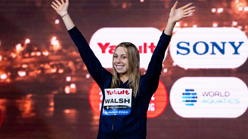 En idrottare sträcker upp sina armar i luften som en gest av seger eller glädje, iförd en mörk outfit med sponsorlogotyper, inklusive ”Yogurt” och ”Walsh.” Bakgrunden visar suddiga ljus och logotyper för företag relaterade till evenemanget.