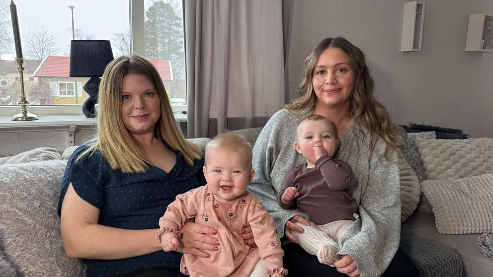Två kvinnor sitter tillsammans i en vardagsrumsmiljö med fönster i bakgrunden. Den ena kvinnan håller en baby i sitt knä, medan den andra kvinnan också har en baby sittande i knät. Rummet har mjuka färger och ljus inredning som ger en hemtrevlig känsla.