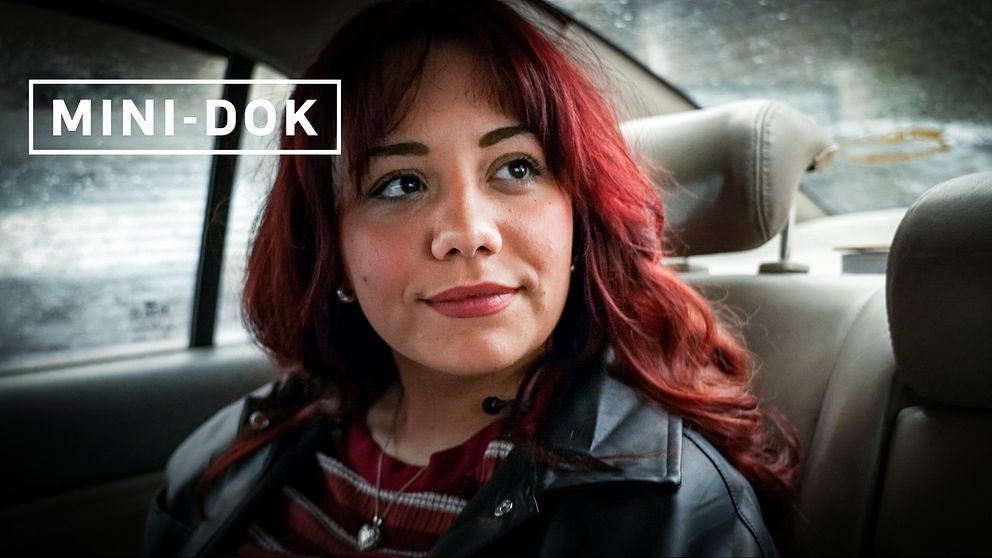 Person med rött hår sitter i baksätet av en bil, märkt ”MINI-DOK”. Hon tittar snett uppåt förbi kameran och hon ler lätt.