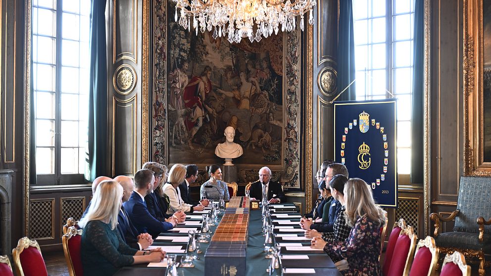 Kung Carl XVI Gustaf leder ett formellt möte vid ett stort konferensbord i ett elegant rum med kristallkrona och väggbonad. Flera personer, både män och kvinnor, sitter runt bordet.