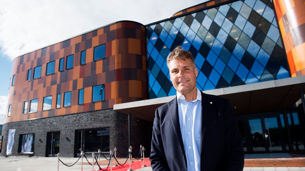 En person står framför en modern byggnad med en fasad i orange och blått glas, som präglas av geometriska mönster. Röda mattan är utrullad och avspärrningar finns i förgrunden, vilket tyder på en VIP-händelse eller invigning.