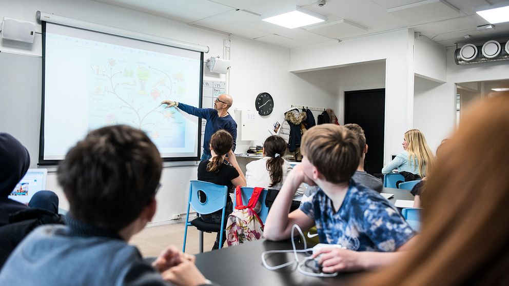Manlig lärare står och pekar på en projicerad bild längst fram i ett klassrum, med barnens blickar riktade framåt.