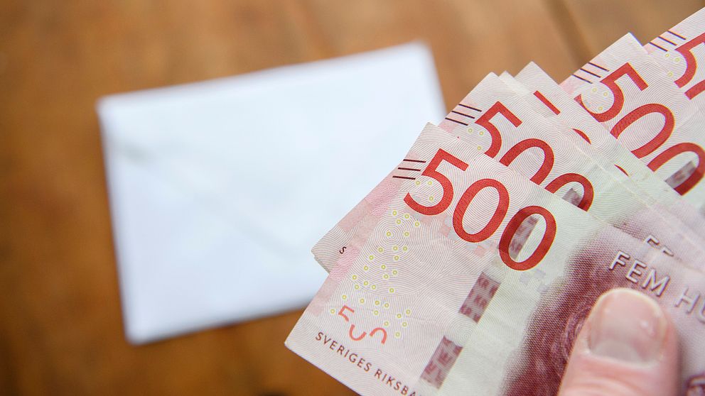 En hand håller flera svenska 500-kronors sedlar, med ett vitt kuvert i bakgrunden på en träytan.