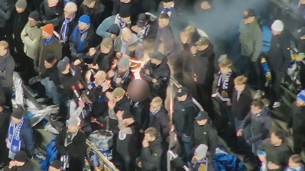 Folksamling klädd i mörka kläder, med någon hållande en fackla, omgiven av rökmoln.