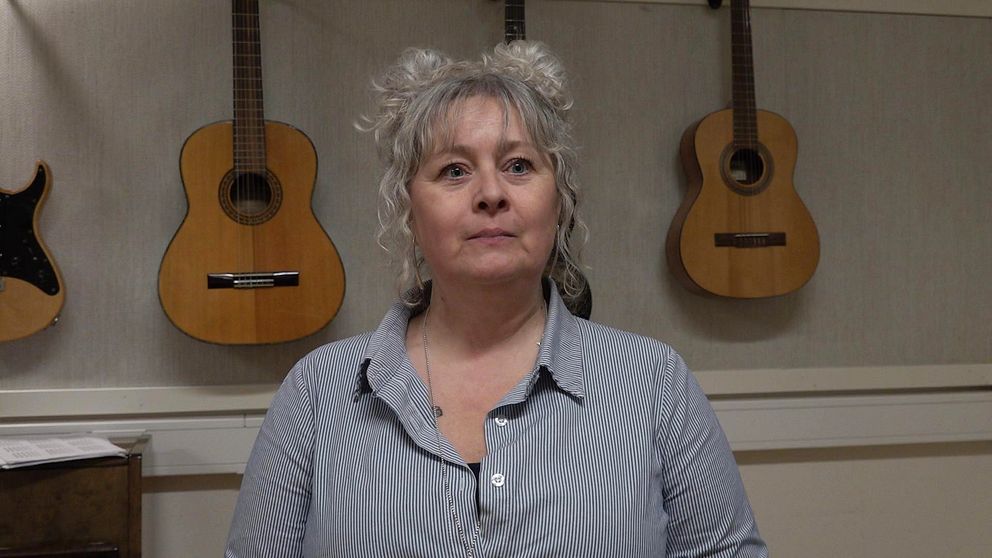 En kvinna sitter och ser sammanbiten ut, bakom hänger flera gitarrer.