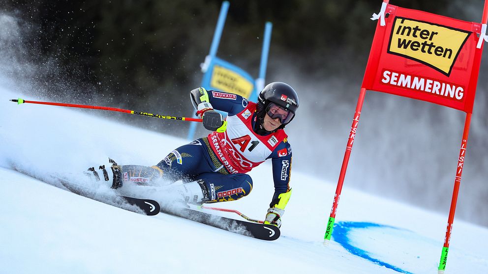 En alpinskidåkare utför en sväng på en snötäckt backe, passerar genom en port markerad med en flagga som har texten ”interwetten SEMMERING”. Skidåkaren är iförd en färgglad tävlingsdräkt och har skidor och störtskydd. Snön sprutar upp bakom dem i en sportig och dynamisk scen.