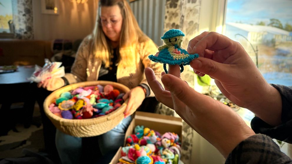 En person håller upp en färgglad virkade figur med en grön och blå klänning. I bakgrunden sitter en annan person med en stor korg fylld med fler virkade figurer i olika färger och former. Rummet är ljust med fönster mot en naturlandskap.
