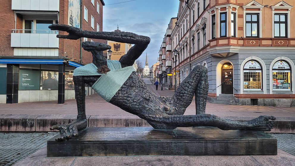 Skulptur av en liggande figur i en stadsmiljö, som pekar åt vänster och bär en ljus tröja. Omgiven av byggnader och gator med affärer i bakgrunden.