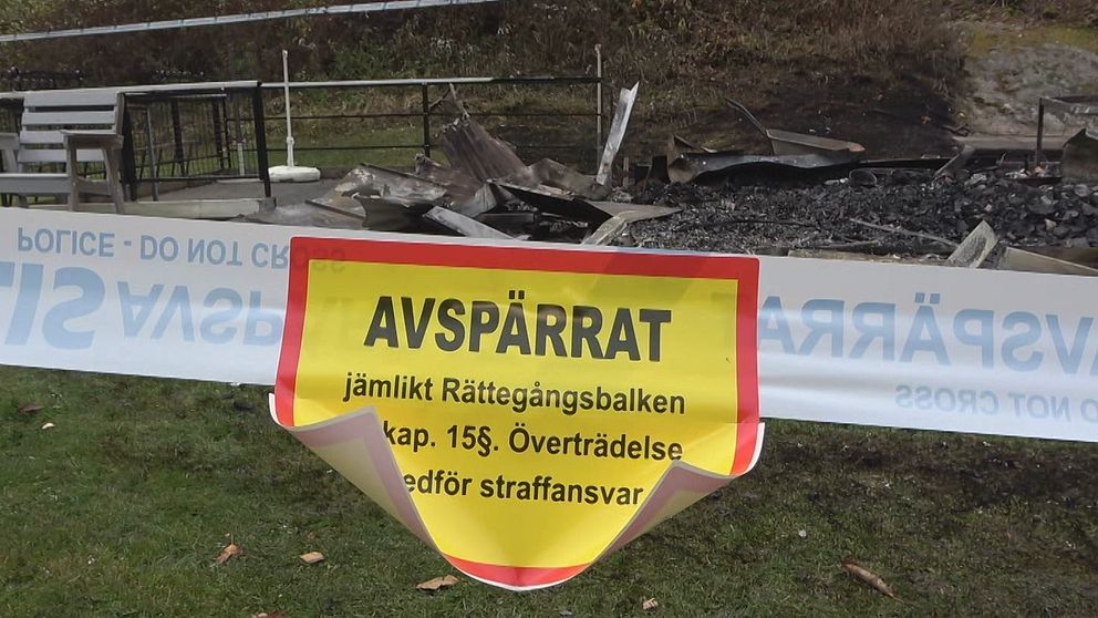 Avspärrat band med en tydlig varningstext som informerar om att området är stängt enligt Rättegångsbalken, i bakgrunden syns förbrända rester av möbler och en askaområde.
