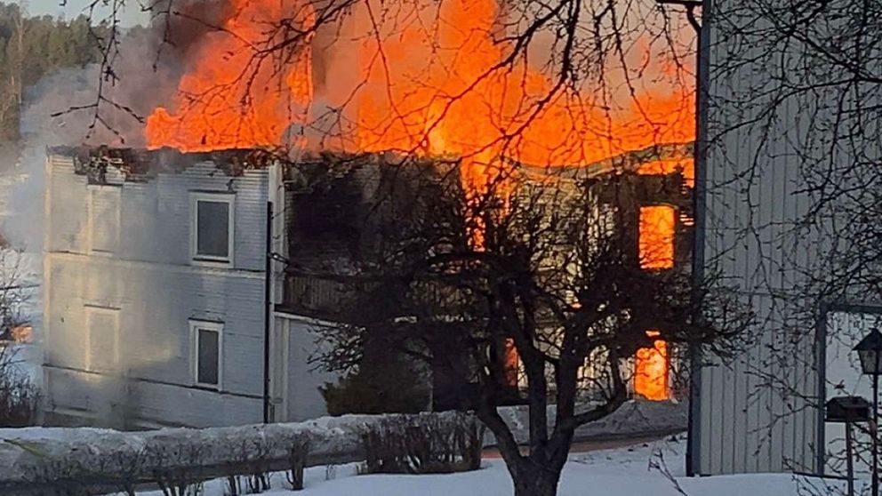 En byggnad är helt övertänd av lågor, med intensiv rök och eld som slår ut från taket. Snö täcker marken runt omkring, vilket kontrasterar mot elden.