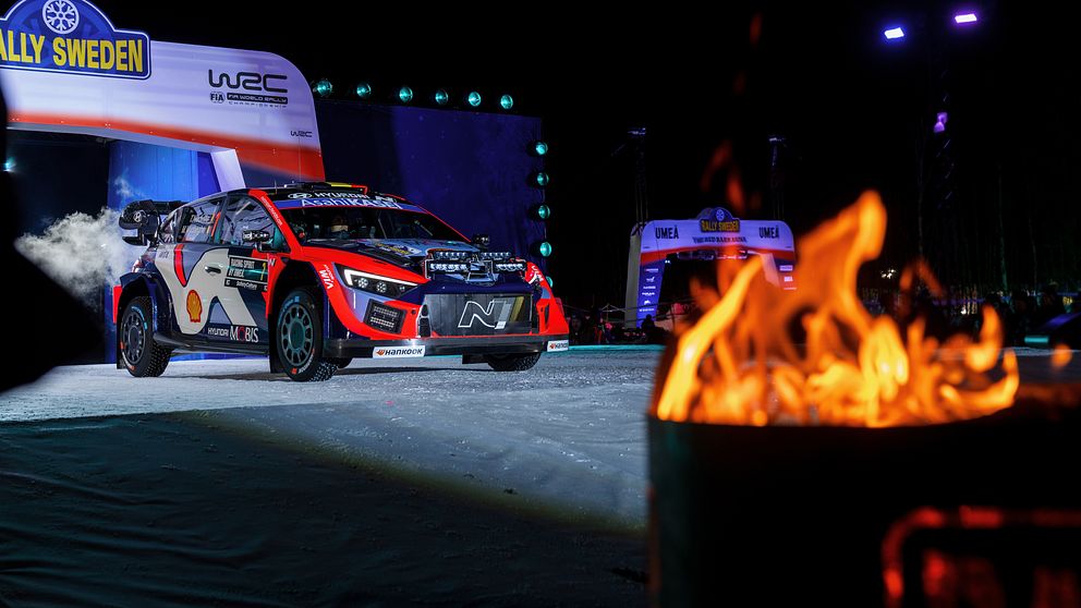 En rallybil deltar i ett nattligt event under Rally Sweden, med ett eldsken i förgrunden som skapar dramatisk belysning. Bakgrunden visar en belyst scen med skyltar och ytterligare en rallybil.