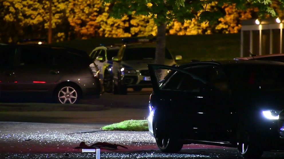 Nattbild av en upplyst parkering där en svart bil med öppen dörr syns i förgrunden. Glassplitter ligger på marken i närheten av bilen. I bakgrunden syns fler parkerade bilar och ett träd med belysning.