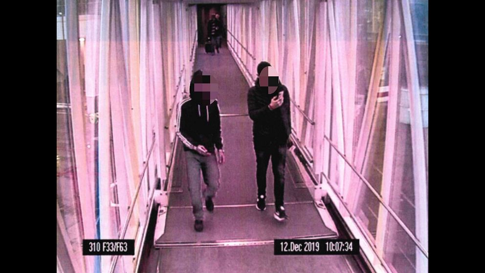 Två män går igenom en korridor på en flygplats, fotograferade av en bevakningskamera.