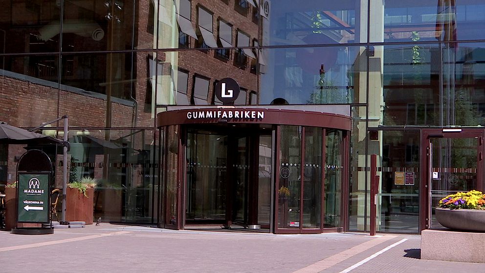 Entrén till Gummifabriken, en modern byggnad med stora glasytor och en roterande dörr, omgiven av stadsmiljö och blomsterarrangemang.