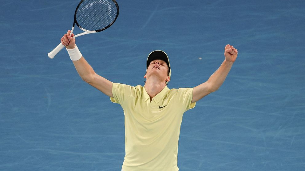 En tennisspelare iklädd ljusgul tröja och vit keps firar en seger på tennisbanan med uppsträckta armar, medan han håller i ett tennisracket.